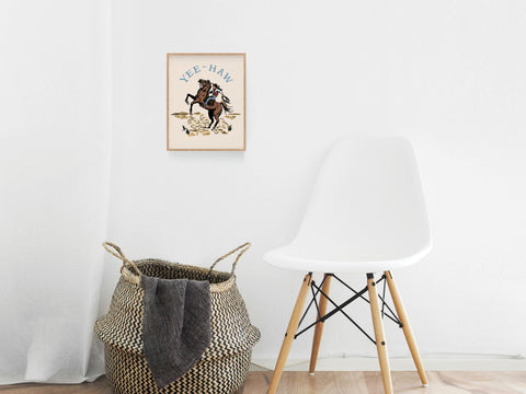 A white chair sits by a woven basket with a blanket. Above, the Yeehaw Cowgirl Art Print by Onderkast Studio, featuring a horse and “Yee-Haw,” brings country flair to the bright, minimal space.