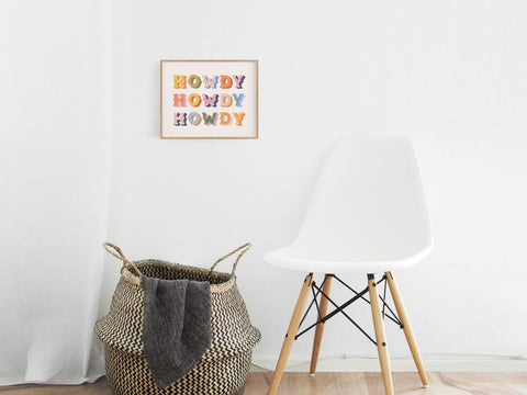 A modern, minimalist room features a white chair, a woven basket with a blanket, and the Howdy Howdy Howdy Art Print by Onderkast Studio on the wall, adding playful cowboy charm with its colorful text design.