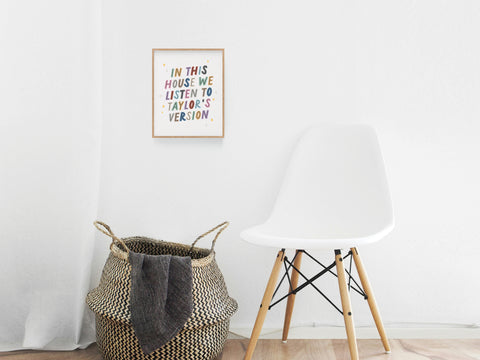 A white chair sits beside a woven basket with a blanket. Above, the Taylor’s Version Art Print by Onderkast Studio hangs on the wall, declaring Swiftie pride with its colorful design.