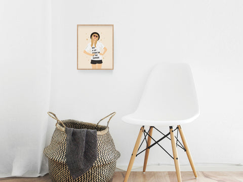 A white modern chair sits by a woven basket in a minimalist room, with the TS Red Art Print by Onderkast Studio—album art–inspired and featuring a woman in sunglasses and a white shirt—hanging on the wall.