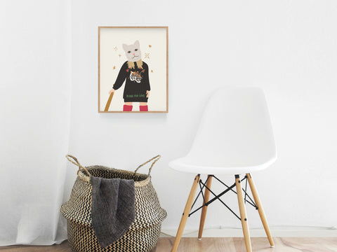 A minimalistic room with a white chair, a woven basket with a gray towel, and Onderkast Studios TS Reputation Art Print on the wall—showing cat mask art of a character holding a bat in black sweater and red pants.