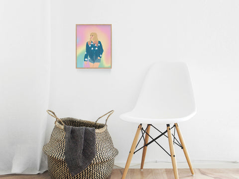 A modern interior with a white chair, woven basket, and the Onderkast Studio TS Lover Art Print—vividly printed, it features a girl in a blue sweater on a pastel rainbow background, making colors glow against the white wall.