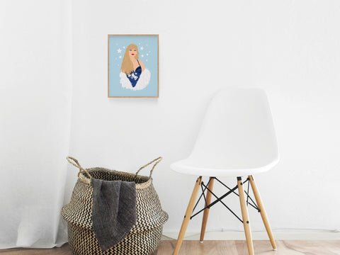 A modern minimalist room with a white chair, woven basket, and Onderkast Studios TS Midnights Art Print—a framed illustration of a blonde woman in a starry blue outfit—adds style to the light blue wall.