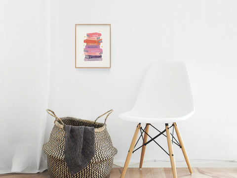 A modern minimalist room features a white chair, woven basket, and the Smut Book Stack Art Print by Onderkast Studio on the wall. Light wood floors and soft natural light add to the clean, cozy Bookish Spaces vibe.