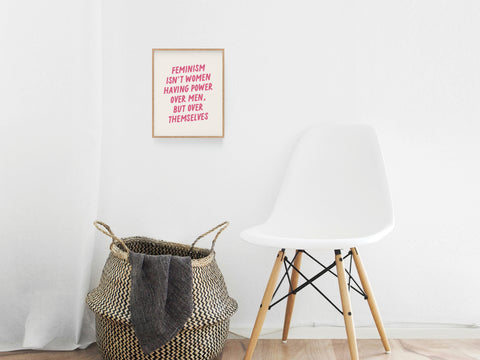 A white chair sits next to a woven basket with a blanket, set against a white wall. Above, the Feminism Art Print by Onderkast Studio, featuring pink text, adds female empowerment to the minimalist decor.