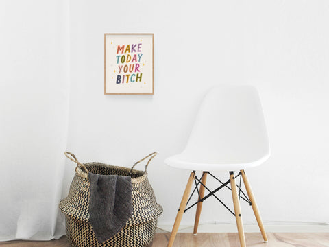 A minimalist room featuring a white chair, woven basket with black towel, and the Make Today Your Bitch Art Print from Onderkast Studio—an inspiring, colorful statement for your home workspace.