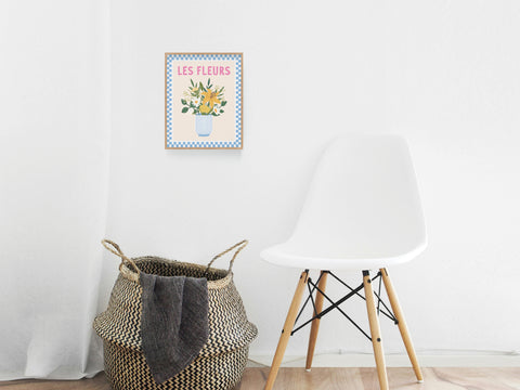 A white modern chair sits beside a woven basket holding a blanket. Above, the Les Fleurs Art Print by Onderkast Studio brings Parisian charm to the white wall with its elegant vase of flowers and stylish LES FLEURS lettering.