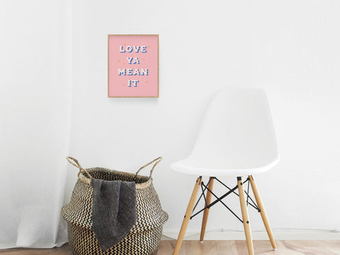 A woven basket and a white modern chair sit by a wall featuring Onderkast Studios Love Ya Mean It Art Print in a pink frame. The space is minimalistic and bright, accentuating the playful, premium vibe of the artwork.