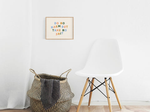 A minimalist room features a white chair, a woven basket with a blanket, and Onderkast Studio’s Do No Harm Art Print on the wall, displaying “Do no harm but take no shit” in vibrant colors for a premium touch.