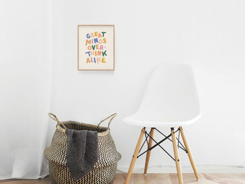 A white modern chair sits by a woven basket and the vibrant Great Minds Overthink Alike Art Print by Onderkast Studio, creating a colorful office vibe with white walls and light wood floors.