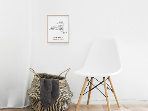 A white chair beside a woven basket with a gray blanket sits below the Onderkast Studio New York Map Art Print, which hangs on a plain white wall and brings a premium touch to the minimalist room.