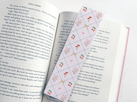 An open book holds an Easyhand Goods Coquette Pattern Laminated Bookmark, featuring bows, mushrooms, flowers, and strawberries in pastel colors. The bookmark’s design is visible on the book’s pages. Printed in the USA.
