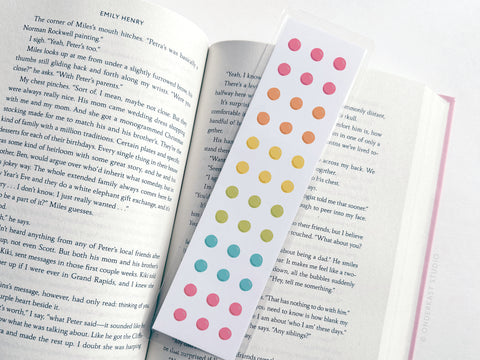 A white Button Candy Laminated Bookmark from Easyhand Goods, featuring vibrant polka dots, lies on open book pages with visible black text. The bookmark and book edges are clearly shown.