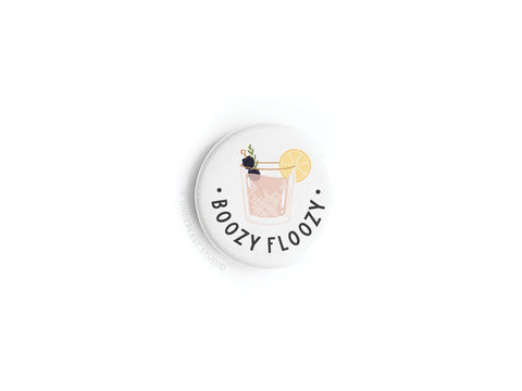 The Boozy Floozy Cocktail Button Magnet by Onderkast Studio features a white round design with a cocktail illustration, lemon slice, flower garnish, and bold “BOOZY FLOOZY” text.