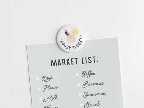 A market list is held on a white surface by the Boozy Floozy Cocktail Button Magnet from Onderkast Studio, which features spirited sass—a lemon slice and bold “BOOZY FLOOZY” text. The list includes eggs, flour, milk, coffee, bananas, cinnamon, and bread.
