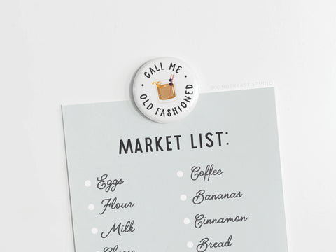 The Call Me Old Fashioned Button Magnet from Onderkast Studio, featuring a classic cocktail design, keeps your shopping list—eggs, flour, milk, coffee, bananas, cinnamon, and bread—secure with vintage charm.