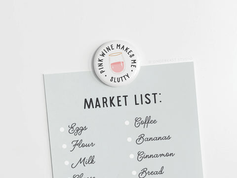 The Pink Wine Makes Me Slutty Button Magnet by Onderkast Studio secures a grocery list—including eggs, flour, milk, coffee, bananas, cinnamon, and bread—on a white surface for wine lovers.