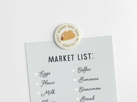 A grocery list is held on a white surface with the Onderkast Studio “Don’t Wanna Taco ‘Bout It” Button Magnet, featuring a taco and witty phrase—perfect playful kitchen decor for taco lovers.