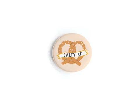The Onderkast Studio Salty AF Pretzel Button Magnet showcases two salted pretzels and a bold yellow SALTY AF banner on a beige background—perfect for snack lovers.