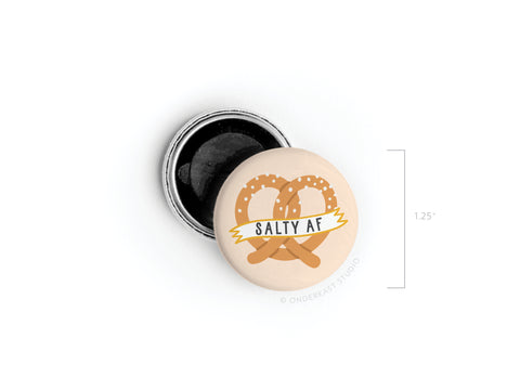Show your snack love with Onderkast Studios Salty AF Pretzel Button Magnet—a 1.25-inch round accessory featuring a bold pretzel design, perfect for adding fun flair to any space.