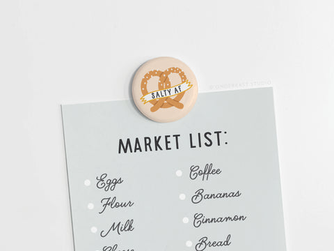 A market list in black cursive is held by Onderkast Studio’s Salty AF Pretzel Button Magnet—perfect for snack lovers. The list includes eggs, flour, milk, cheese, coffee, bananas, cinnamon, and bread.