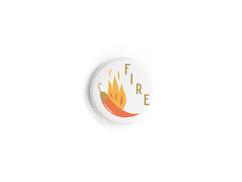 The Onderkast Studio Fire Chili Button Magnet is a round white magnet with a bold chili pepper design, vibrant red and yellow flames, and the word FIRE in gold letters displayed vertically next to the flames.