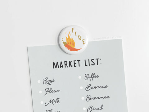 The Fire Chili Button Magnet by Onderkast Studio features a bold chili pepper and flame design labeled FIRE, attached to a market list with eggs, flour, milk, coffee, bananas, cinnamon, and bread.