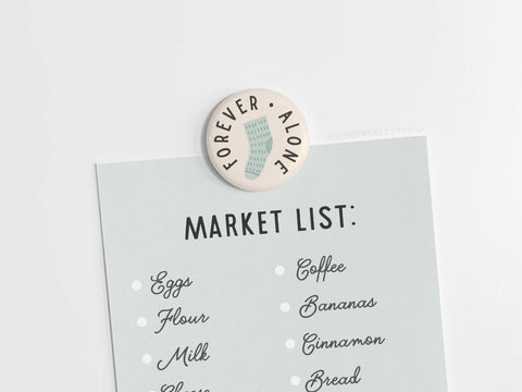 The Forever Alone Sock Button Magnet by Onderkast Studio adds playful style as it holds a market list—eggs, flour, milk, cheese, coffee, bananas, cinnamon, and bread—to any white surface.