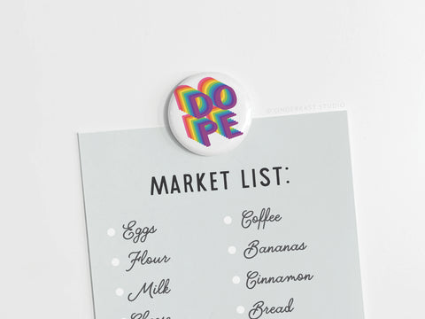 A grocery list is secured by the DOPE Button Magnet from Onderkast Studio, featuring bold, colorful 3D letters. High-quality design stands out with eggs, flour, milk, coffee, bananas, cinnamon, and bread on a light background.