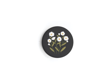 The Onderkast Studio Daisies Button Magnet is a round black magnet featuring a vibrant print of five white daisies with yellow centers and green leaves arranged in a cluster.