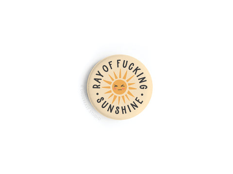 The Ray of Fucking Sunshine Button Magnet by Onderkast Studio is a round beige magnet with a stylized sun, neutral face, and “RAY OF FUCKING SUNSHINE” in bold black letters, vibrantly printed on a white background.