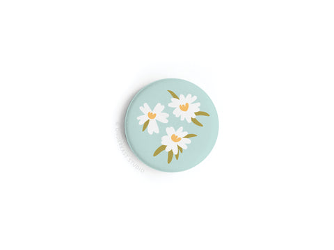 The White Flowers Button Magnet by Onderkast Studio is a round, light blue magnet with an illustration of three white flowers, yellow centers, and green leaves on a plain white background.