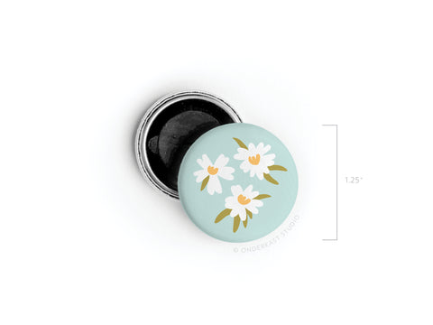 A small round tin, 1.25 in diameter, with a light blue lid featuring Onderkast Studio’s White Flowers Button Magnet design—three simple white daisies. The tin is open, showing a dark interior.