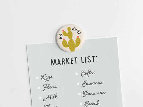 The No Hugs Cactus Button Magnet by Onderkast Studio holds a blue market list with eggs, flour, milk, coffee, bananas, cinnamon, and bread—a quirky magnet perfect for introverts.