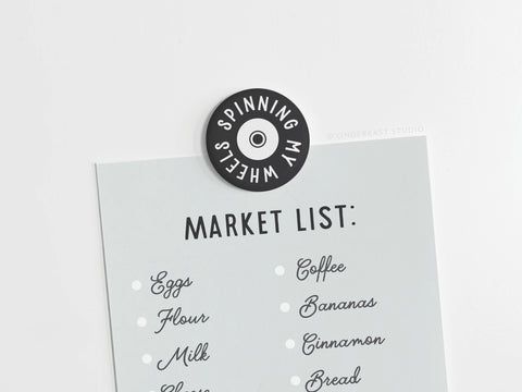 A grocery list is held on a white surface with the discontinued Smagnetning My Wheels Button Magnet by Onderkast Studio, featuring vibrant colors. Items listed: eggs, flour, milk, cheese, coffee, bananas, cinnamon, and bread.