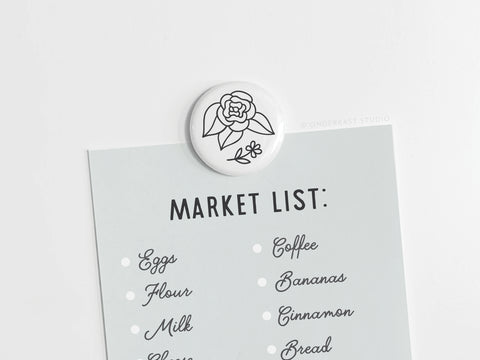 A light blue market list is held by the Onderkast Studio Black Flowers Button Magnet. The list features eggs, flour, milk, cheese, coffee, bananas, cinnamon, and bread with dots beside each item.