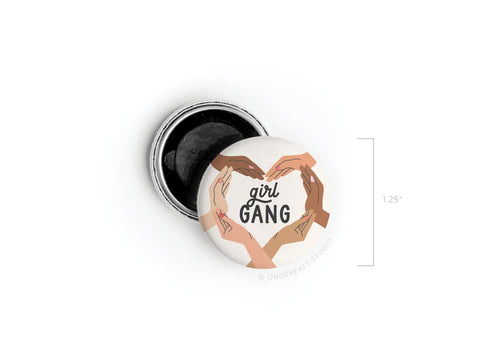 The Girl Gang Button Magnet by Onderkast Studio features hands of different skin tones forming hearts around “girl GANG,” symbolizing female friendship. The round, 1.25-inch magnet is shown open with its black back visible.