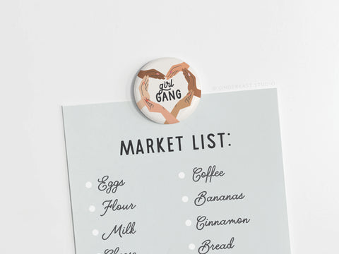A market list is held by the Girl Gang Button Magnet from Onderkast Studio, featuring diverse hands forming a heart around girl gang. It celebrates friendship and lists eggs, flour, milk, cheese, coffee, bananas, cinnamon, and bread.