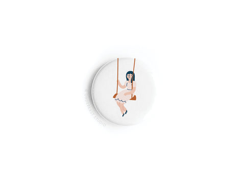 The Girl on a Swing Button Magnet by Onderkast Studio features a nostalgic illustration of a blue-haired woman in a white dress sitting gracefully, set against a plain white background.
