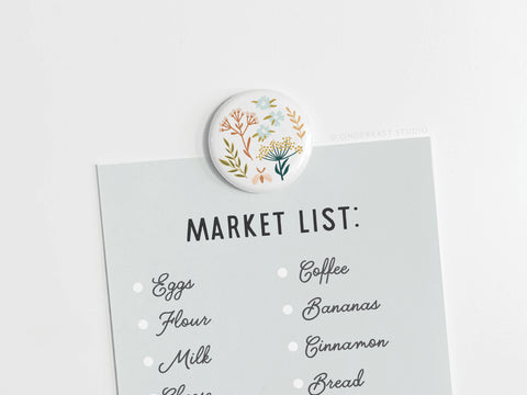The Onderkast Studio Soft Florals Button Magnet holds a pale blue Market List—featuring eggs, flour, milk, coffee, bananas, cinnamon, and bread—on a white surface.