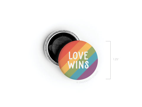 The Love Wins Button Magnet by onderkast studio features rainbow stripes and LOVE WINS in white text. This 1.25-inch button magnet supports LGBTQ+ rights and is shown partially open, revealing its black interior.