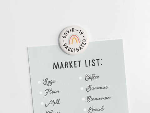 A Market List is attached to a white surface with an eye-catching Onderkast Studio Covid-19 Vaccinated Button Magnet (DISCONTINUED), featuring a rainbow. Items listed include eggs, flour, milk, coffee, and bananas.