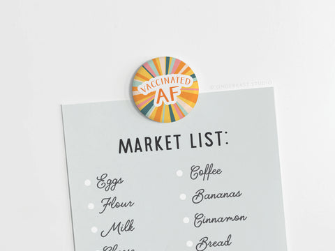 A brightly printed Vaccinated AF Button Magnet by Onderkast Studio (DISCONTINUED) holds a grocery list—eggs, flour, milk, cheese, coffee, bananas, cinnamon, bread—showcasing its bold colors and quality.