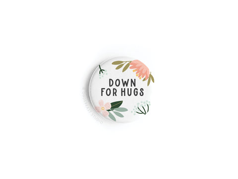 The Down For Hugs Button Magnet by Onderkast Studio features black text, vibrant pink and yellow flowers, and green leaves on a white background.