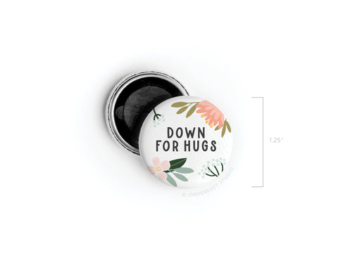 The Down For Hugs Button Magnet by Onderkast Studio features vibrant pink and yellow flowers with “DOWN FOR HUGS” in bold black letters. Shown open to reveal its black interior, it measures 1.25 inches in diameter.