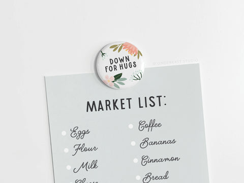 A brightly colored Down For Hugs Button Magnet by Onderkast Studio secures a pale blue market list—eggs, flour, milk, cheese, coffee, bananas, cinnamon, and bread—on a white surface.
