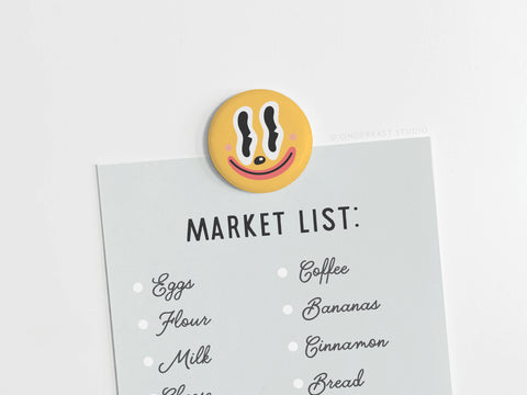 A shopping list with eggs, flour, milk, coffee, bananas, cinnamon, and bread is held by the Trippy Happy Face Button Magnet from Onderkast Studio—its bold black and white eyes and red mouth on yellow give a playful psychedelic vibe.
