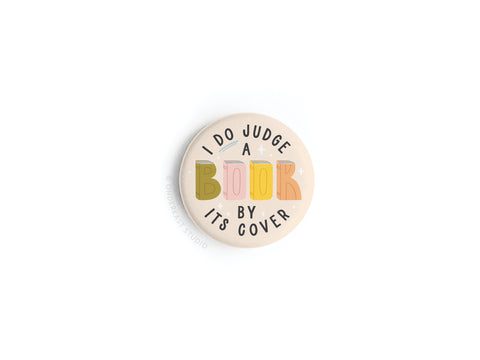 The I DO Judge a Book By Its Cover Button Magnet by Onderkast Studio features playful, multicolored text on a light background—an eye-catching and fun magnet for any collection.
