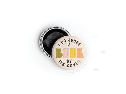 The Onderkast Studio I DO Judge a Book By Its Cover Button Magnet features colorful, playful lettering and comes with a 1.25-inch diameter for scale, shown next to an open pin backing.