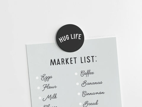 A market list featuring eggs, flour, milk, cheese, coffee, bananas, cinnamon, and bread in a stylish font is clipped with the Onderkast Studio Hug Life Button Magnet—a cozy touch in vibrant prints.